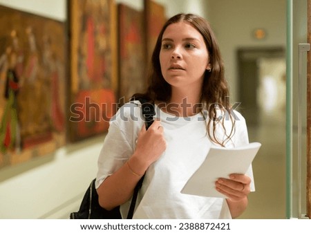 Portrait of a focused girl standing in the museum hall next to an exhibit located in a glass cabinet, with a booklet of the ..exhibition program Royalty-Free Stock Photo #2388872421
