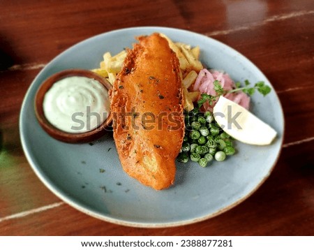 Fish and chips using dory fish accompanied by fried potatoes, vegetables and tartar sauce on a white plate on a brown wooden table