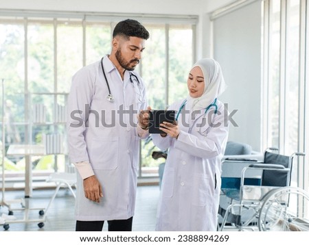 Asian Indian professional successful bearded male doctor in white lab coat uniform with stethoscope standing talking discuss brainstorm with female muslim colleague in hijab holding tablet computer.
