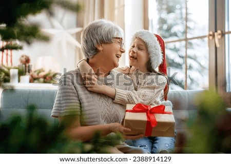 Merry Christmas and Happy Holidays! Cheerful grandma and her cute grand daughter girl exchanging gifts. Granny and little child having fun near tree indoors.