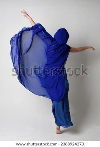 Full length portrait of beautiful young female model elegant blue fantasy  ball gown and flowing cape with hood.
Standing pose, walking away facing backwards. Isolated on whit studio background.