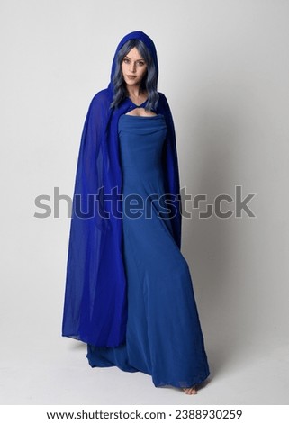 Full length portrait of beautiful female model wearing elegant fantasy blue ball gown and flowing cape with hood.
Standing pose, with gestural arms reaching out . Isolated on white studio background.