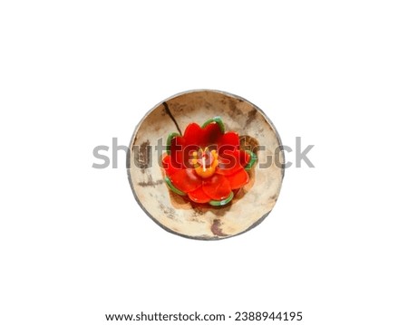 In the picture is a red and green candle in the shape of a lotus with yellow stamens and a white light string in the center of the picture, placed in a brown-striped coconut shell.