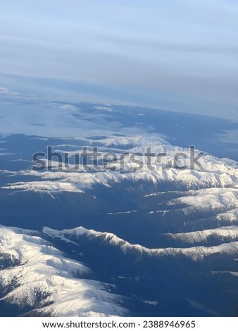 From the airplane, a mountain veiled in a glistening blanket of snow. The peaks stand resolute, their icy ridges contrasting against the azure sky.