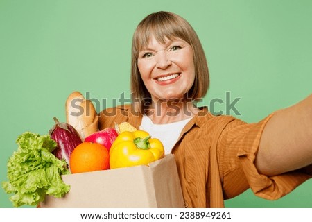 Close up elderly woman wear brown shirt casual clothes hold shopping paper bag with food products do selfie shot pov on mobile cell phone isolated on plain green background. Delivery service from shop