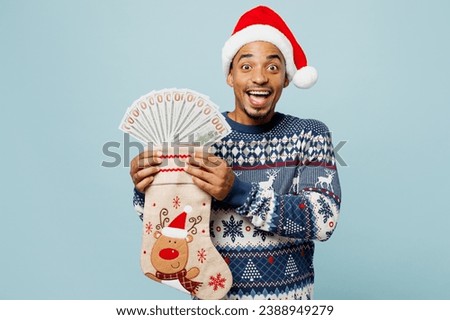 Young fun man wear sweater Santa hat posing hold stocking for gifts fan of cash money in dollar banknotes isolated on plain blue background. Happy New Year 2024 celebration Christmas holiday concept