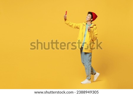 Full body young woman wear waterproof raincoat outerwear red hat do selfie shot on mobile cell phone show v-sign isolated on plain yellow background. Outdoors lifestyle wet fall weather season concept