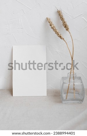 Blank paper card mockup and dried meadow grass spikelets in vase on table with linen neutral beige tablecloth and oat color wall background, aesthetic home interior business brand or wedding template.