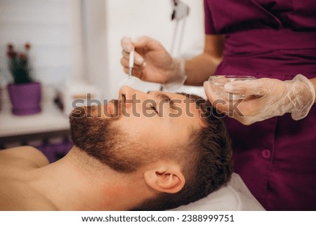 Cosmetologist applying cosmetic mask on man's face in spa salon. Handsome bearded man getting facial skin care treatment. . High quality photo