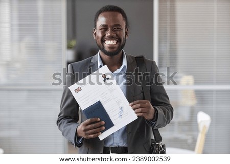 Young cheerful African American male applicant in formalwear looking at camera while showing approved visa application form Royalty-Free Stock Photo #2389002539