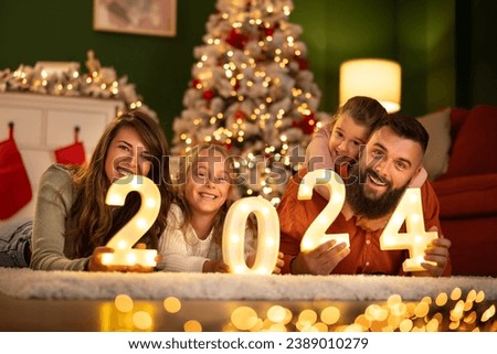 Parents and children having fun celebrating New Year together at home, lying by the Christmas tree holding illuminative numbers 2024 Royalty-Free Stock Photo #2389010279