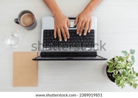 Portrait of a cheerful young woman typing and working on a laptop while being seated next to a table at home. Portrait of a young woman working on a laptop at home. 