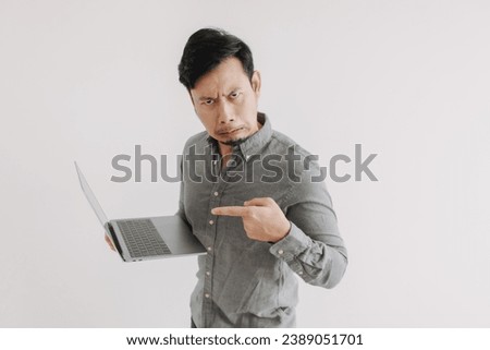 Asian man with beard wear grey shirt, disappointed and unsatisfied face, holding notebook hand pointing at laptop, funny ugly face looking at camera isolated over white background wall.