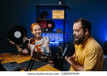 Music bloggers recording a new episode of their podcast. A man and a woman are talking about music in the studio, the woman is holding a vinyl record in her hands.