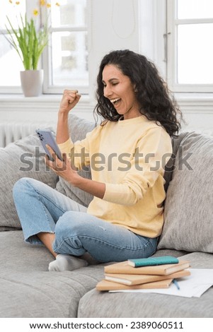Vertical photo of a young happy girl rejoicing, celebrating Victory, holding her fist victoriously up, looking at the phone screen. The student successfully passed the test or exam. Distance Learning. Royalty-Free Stock Photo #2389060511
