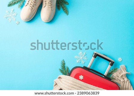 Winter vacation concept. Overhead view of a red suitcase, cozy boots, knitted scarf, fir branches, snowflakes on a bright blue surface, with space for text or adverts