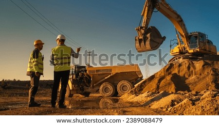 Caucasian Male Real Estate Investor And Project Manager Talking On Construction Site Of Apartment Block. Colleagues Discussing Building Progress. Excavator Loading Sand In Industrial Truck On Warm Day Royalty-Free Stock Photo #2389088479