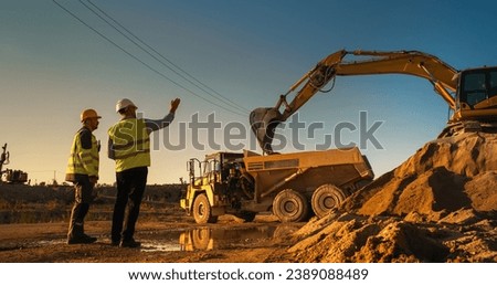 Caucasian Male Real Estate Investor And Civil Engineer Talking On Construction Site Of Apartment Block. Colleagues Discussing Building Progress. Excavator Loading Sand In Industrial Truck On Warm Day