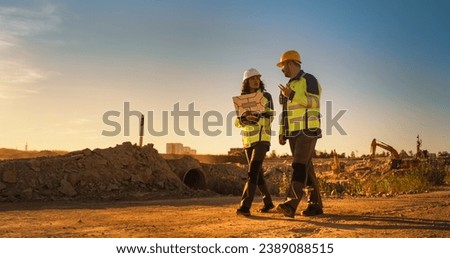 Caucasian Male Architect And Hispanic Female Urban Planner Walking On Construction Site With Laptop Computer And Talking About New Real Estate Project. Construction Equipment Working. Golden Hour