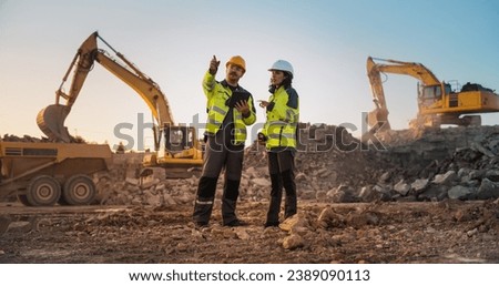 Caucasian Male Civil Engineer Talking To Hispanic Female Inspector And Using Tablet On Construction Site of Apartment Complex. Real Estate Developers Discussing Business, Excavators Working. Royalty-Free Stock Photo #2389090113