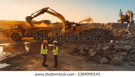 Aerial Drone Shot Of Construction Site On Sunny Evening: Caucasian Male Civil Engineer Talking To Hispanic Female Urban Planner and Using Tablet. Industrial Excavator Loading Rocks Into A Truck.