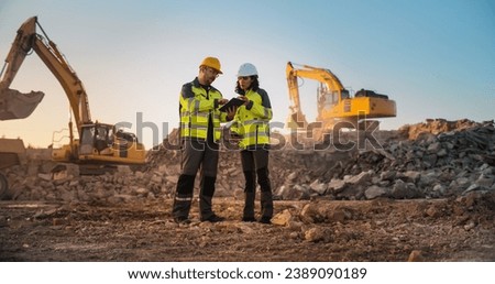 Caucasian Male Civil Engineer Talking To Hispanic Female Inspector And Using Tablet On Construction Site of New Apartment Complex. Real Estate Developers Discussing Business, Excavators Working. Royalty-Free Stock Photo #2389090189