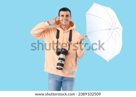 Male photographer with camera and lighting equipment showing "call me" gesture on blue background