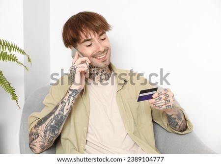 Tattooed young man with credit card talking by mobile phone at home