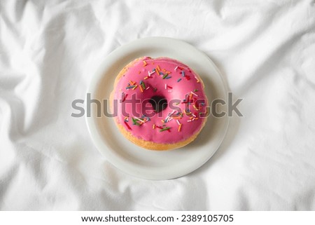 Appetizing pink donut with decorative sprinkles in bed on a white blanket, top view.