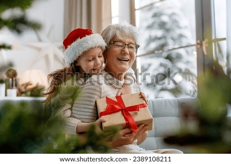 Merry Christmas and Happy Holidays! Cheerful grandma and her cute grand daughter girl exchanging gifts. Granny and little child having fun near tree indoors.