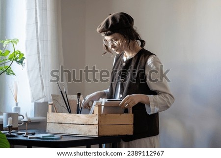 Cartoon artist standing at table, holding wooden box with painting accessories, brushes, sketch books, preparing for work. Workplace of drawing tutor, illustrator creating animation in art school