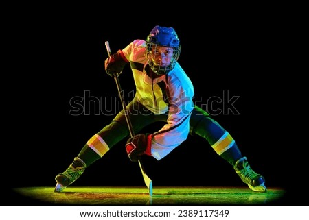 Concentrated young woman, hockey player un uniform and helmet, standing with stick against black studio background in neon light. Concept of professional sport, competition, game, action, hobby