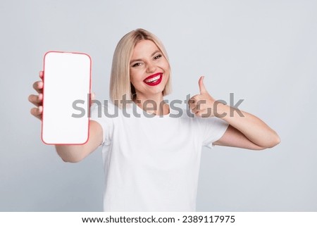 Portrait of toothy beaming girl with bob hairstyle wear white t-shirt show smartphone display thumb up isolated on gray color background