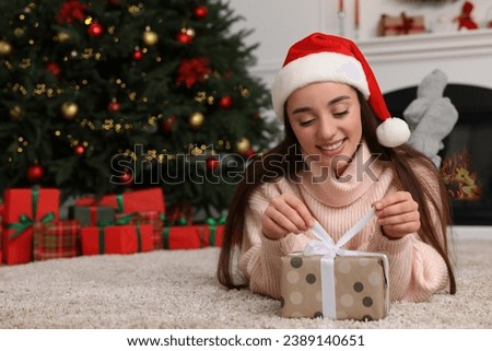 Happy young woman in Santa hat opening gift box in room decorated for Christmas