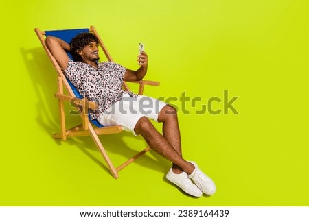 Full body photo of handsome young man hold gadget sit sunbed dressed stylish pink leopard print clothes isolated on yellow color background