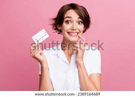 Photo of funny impressed girl with bob hairdo dressed white shirt hold credit card finger on teeth isolated on pink color background