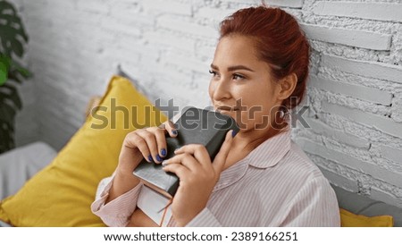 Beautiful young redhead woman, comfortably sitting on bed in her bedroom. morning read holding a book, deep in thought, relaxed and confident at home. Royalty-Free Stock Photo #2389166251