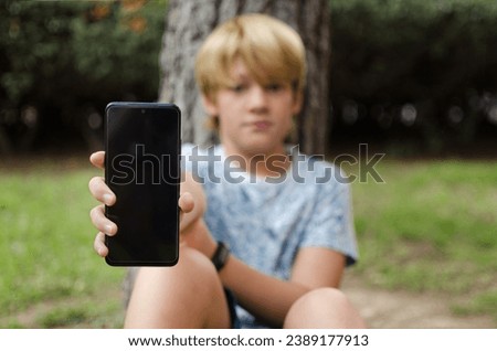 Pretty ten years old blond boy showing mobile phone empty screen. Horizontal photography of a preteen sitting in a park looking at camera. Display mockup mobile app advertisement. Selective focus
