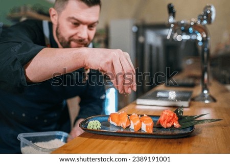 Bearded chef cook dressed black uniform sprinkling the sushi Nigirizushi dish in basement restaurant kitchen. Professional occupation, food preparation, small business and restaurants industry concept Royalty-Free Stock Photo #2389191001