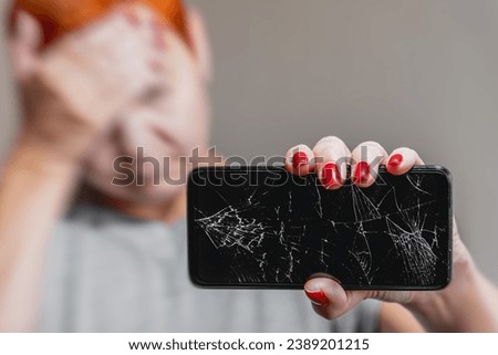 A young woman reacts emotionally to a broken smartphone screen. A pretty woman holds a broken mobile phone in her hand. Selective focus on the broken display of a cracked smartphone. Royalty-Free Stock Photo #2389201215