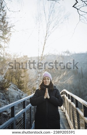 Girl folded hands namaste in front of her chest against the background of the morning sun, pleasant young woman meditating in nature in the cold season. High quality photo