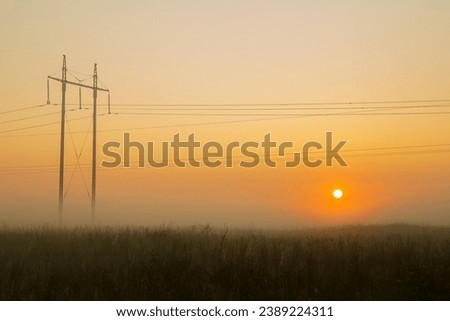 The setting sun in the field against the background of power lines evening dark golden sky and fog. Horizontal photo