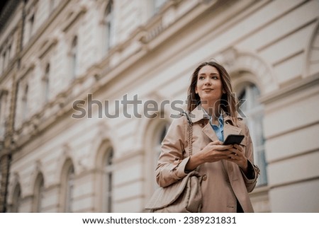 Portrait of a gorgeous blonde smiling woman using mobile phone while outside on a city street. Young female checking her smartphone in an urban business district. Royalty-Free Stock Photo #2389231831