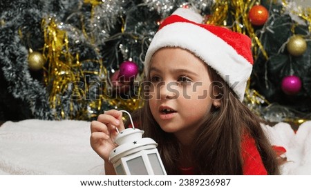 happy girl in santa claus hat hold hand demonstrate x-mas newyear advert promo wear warm winter clothes sings songs, congratulations a New Year's Christmas tree with toys and lights