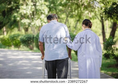 Back view of Asian female doctor helps senior male patient walk to hospital park. Concepts of physicality, caregivers, and therapy. Royalty-Free Stock Photo #2389272267