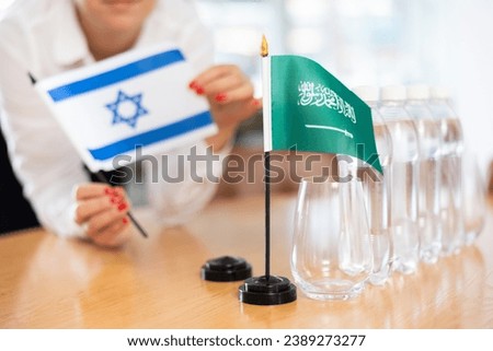 Unrecognizable woman preparing room for international negotiations and communication discussions of leaders. Lady sets miniatures flags of Saudi Arabia and Israel on table. Unfocused shot