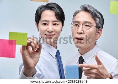Two businessmen are standing on the glass wall of the office, looking at the attached paper and contemplating planning and strategy.