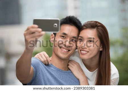 Happy Asian couple using a Smart Phone to take a 'Selie' self portrait. Outdoors in the city.