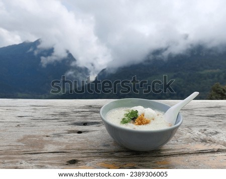 Boiled rice with boiled egg, pork, coriander, and fried garlic in a bowl on the wooden table. Breakfast with beautiful natural landscape in front. Mountains with floating white cloud fog. Thailand.  Royalty-Free Stock Photo #2389306005