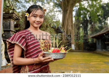 A happy balinese woman in traditional dress with a tray of colorful offerings put some incense sticks 